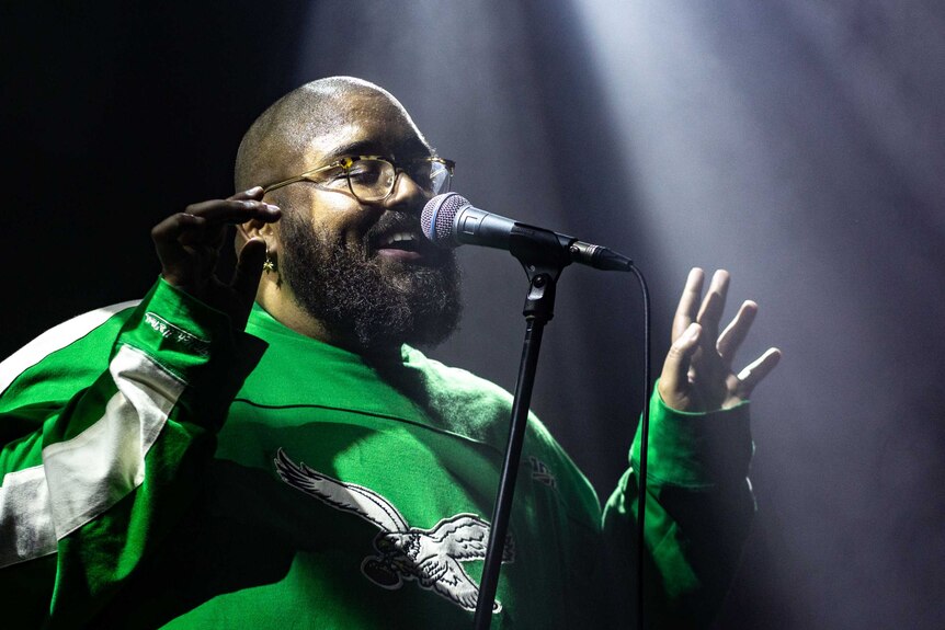 Adrian Eagle wearing glasses and a green jumper, singing into a microphone