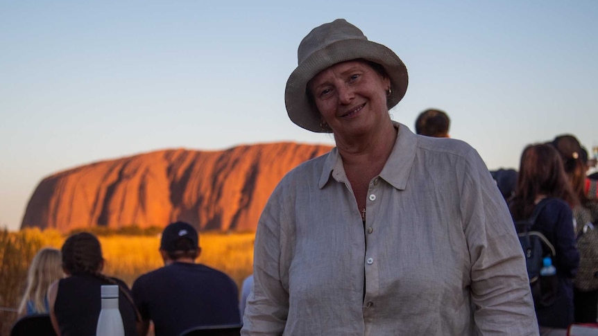 A lady stands in front of a camping table with Uluru in the background