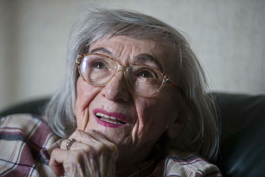And elderly woman stares into the distance with her chin in hand, large horn-rimmed glasses on her face, in her lounge room.