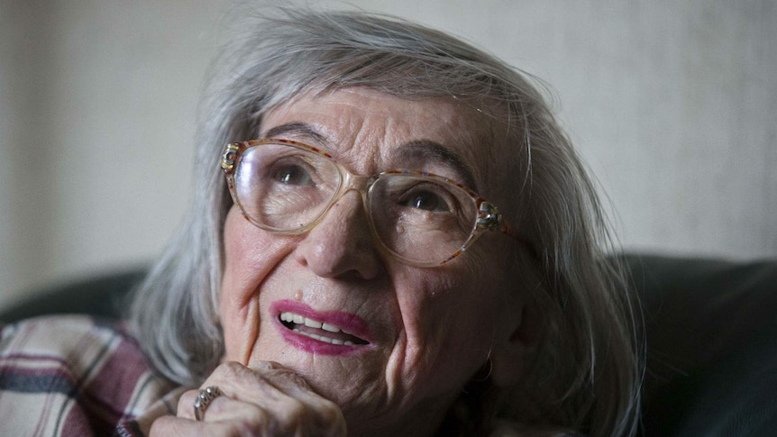 And elderly woman stares into the distance with her chin in hand, large horn-rimmed glasses on her face, in her lounge room.