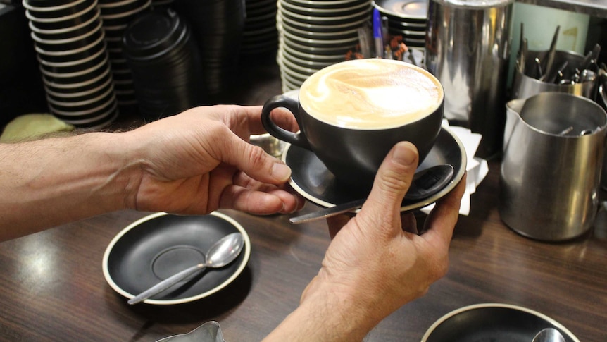 Two hands holding a latte in a black cup and saucer.