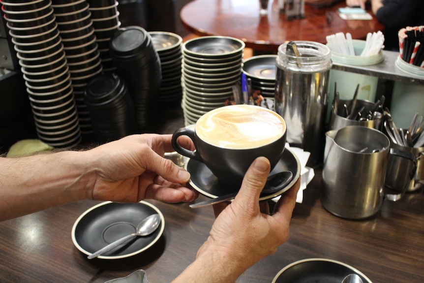 Two hands holding a latte in a black cup and saucer.