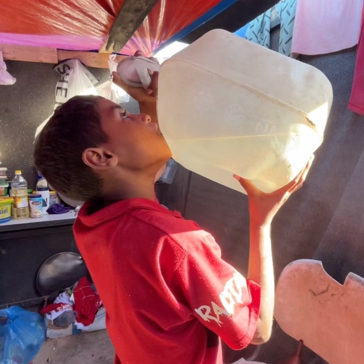 A young boy pictured inside a tent drinking from a large container of water