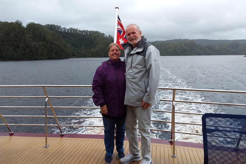 An older man and a woman stand on the deck of a boat with river and mountains behind them.