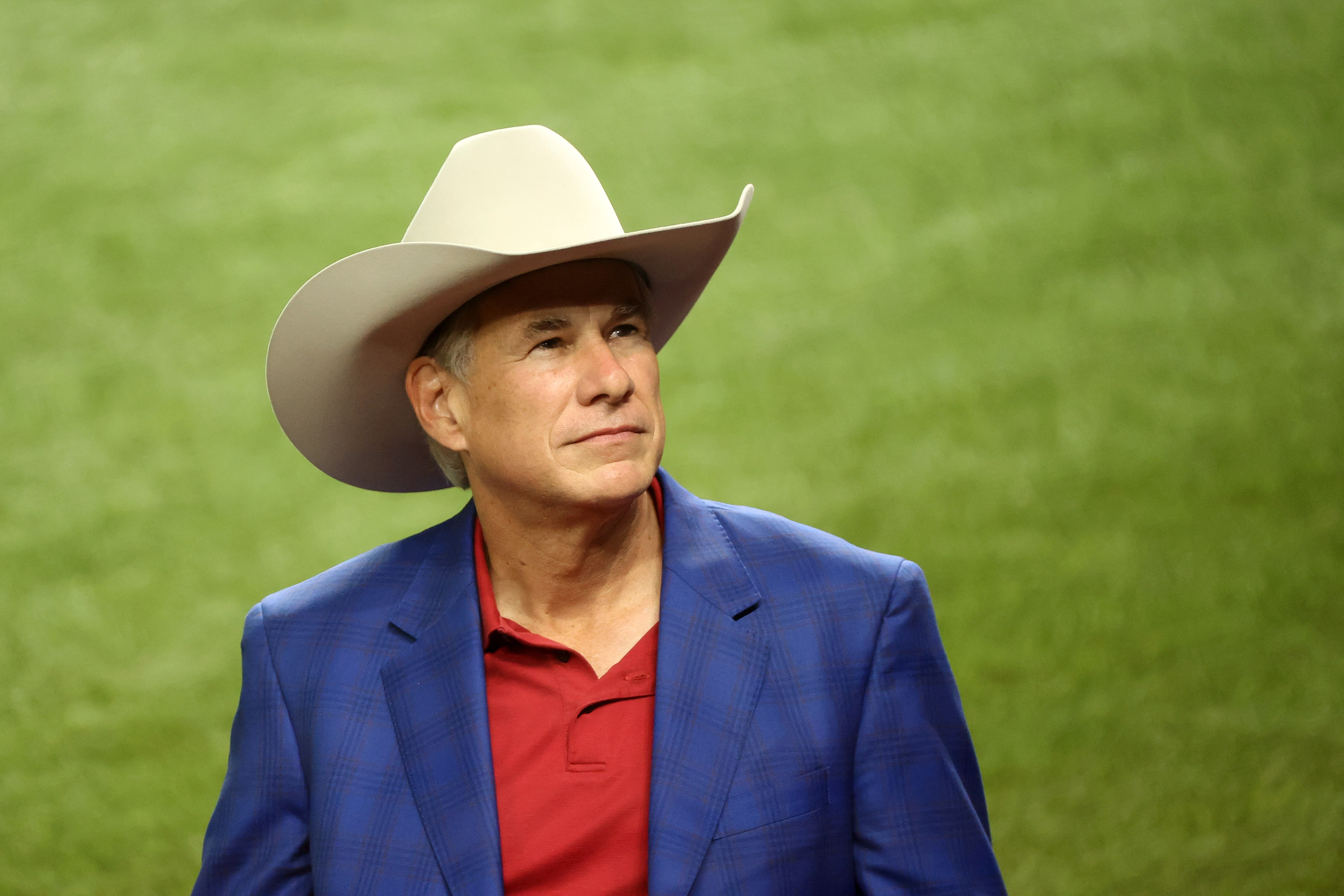 Greg Abbott, wearing a red shirt, blue blazer and large cream Stetson hat, gazes upwards from a green field