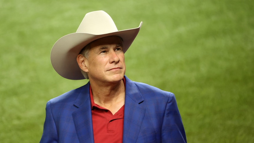 Greg Abbott, wearing a red shirt, blue blazer and large cream Stetson hat, gazes upwards from a green field