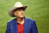 Greg Abbott, wearing a red shirt, blue blazer and large cream Stetson hat, gazes upwards from a green field
