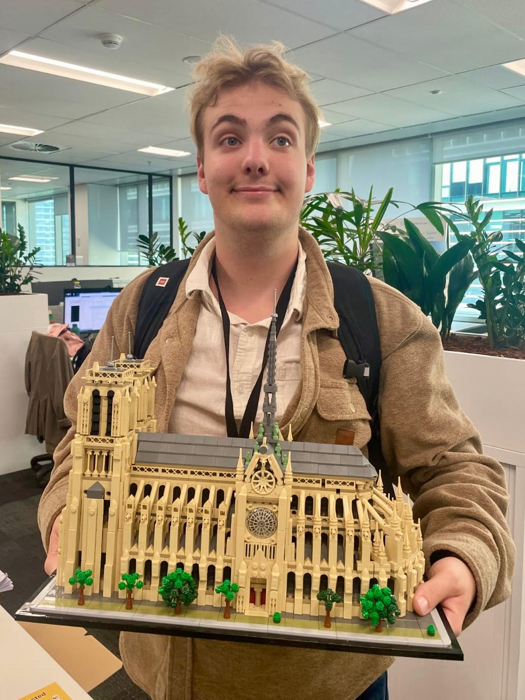 Teenage boy with blond hair wearing white collared shirt, brown jacket, black lanyard and backpack holds replica Lego Notre Dame