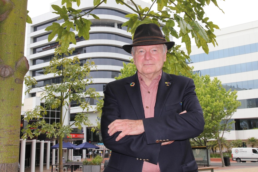 A man in a navy suit and hat stands with arms crossed under a tree