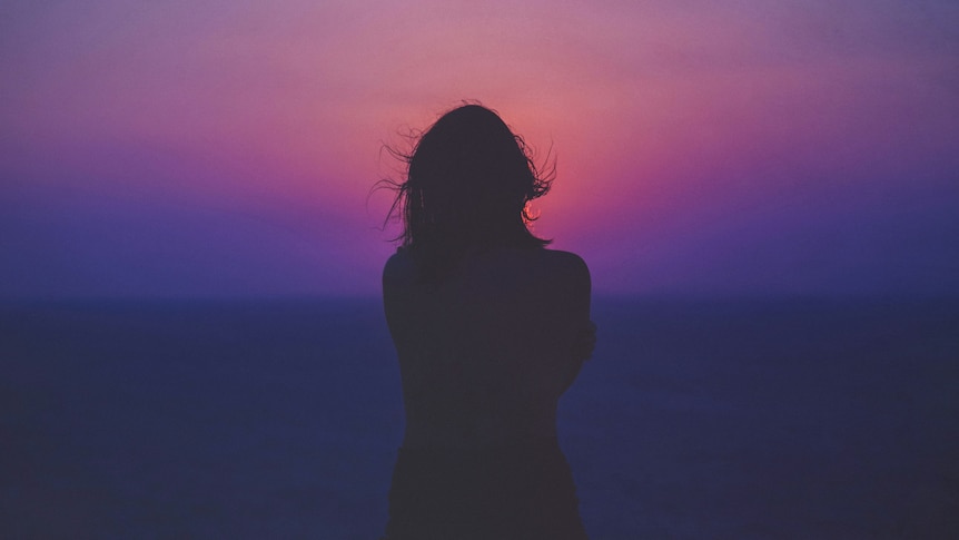 A woman is silouhetted from behind, her hair windblow, as she looks into a bright pink and purple sky.
