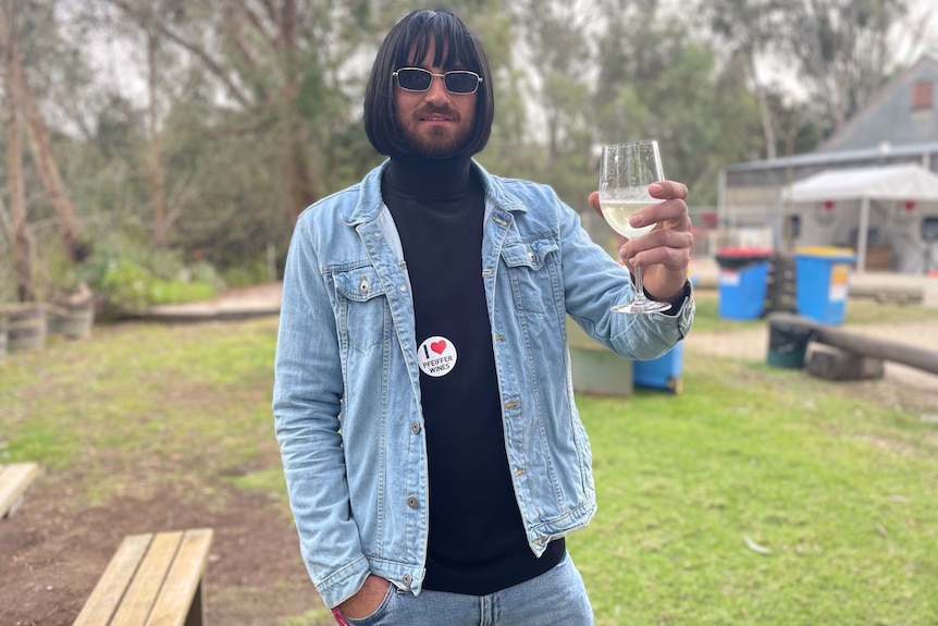 A man dressed in double denim and a wig holds a glass of white wine