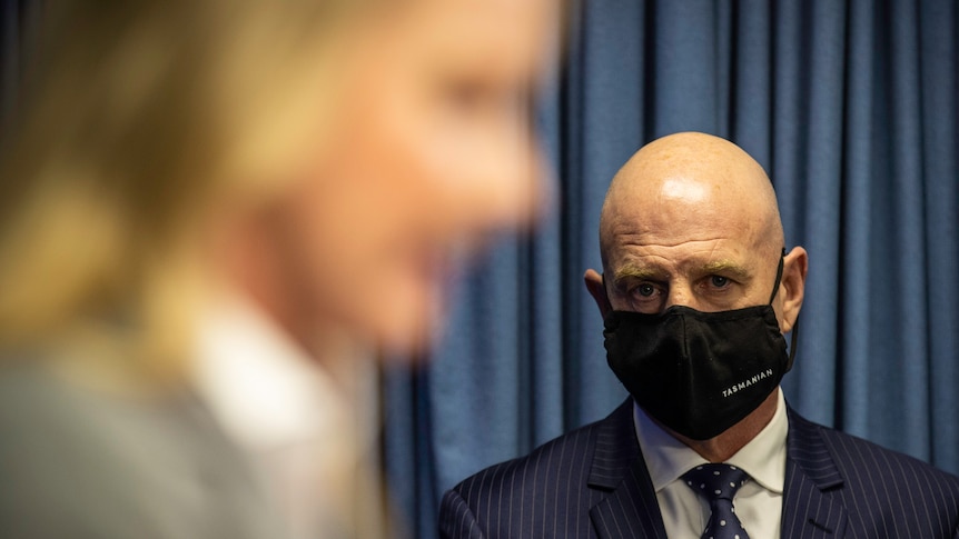 Blurry woman in the foreground watched by a bald man in a face mask.