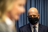 Blurry woman in the foreground watched by a bald man in a face mask.