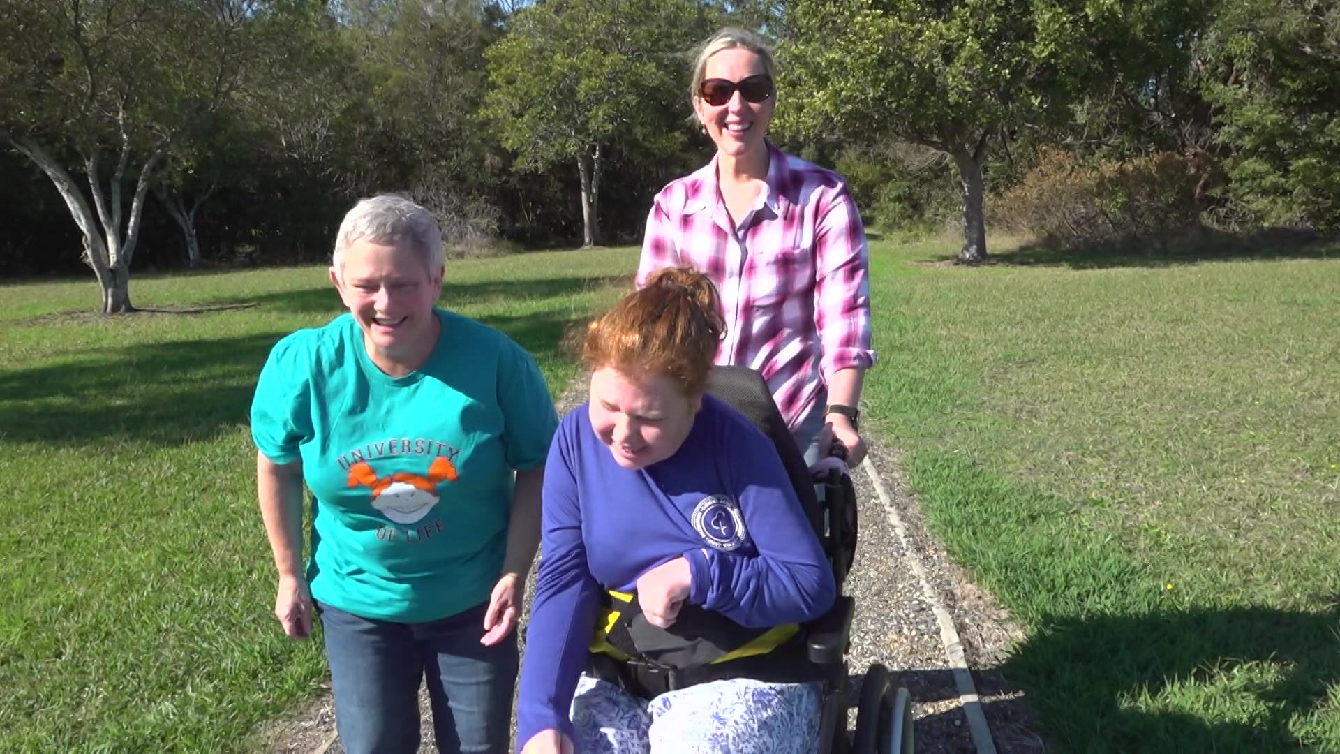 A woman with short grey hair and a blue UOL T-shirt walks beside Lillian and her support worker pushing her wheelchair in a park
