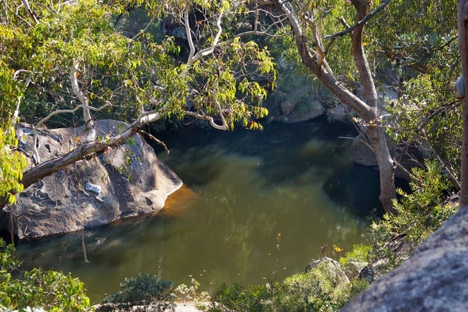 An overhead shot from a rockface shows a calm, still pool of water with trees overhanging