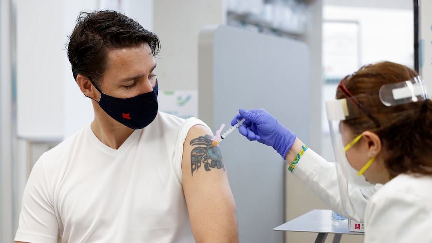 A masked middle aged man with dark hair and a tattoo receives an injection from a masked female nurse at clinic