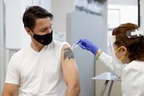 A masked middle aged man with dark hair and a tattoo receives an injection from a masked female nurse at clinic