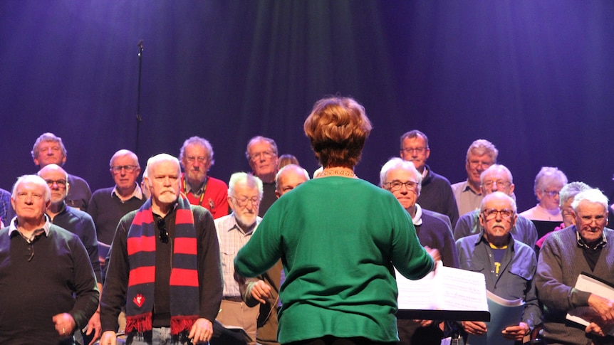 The choir conduct faces away from camera. Her arms are moving to direct the choir in song. Three rows of faces look bacl.