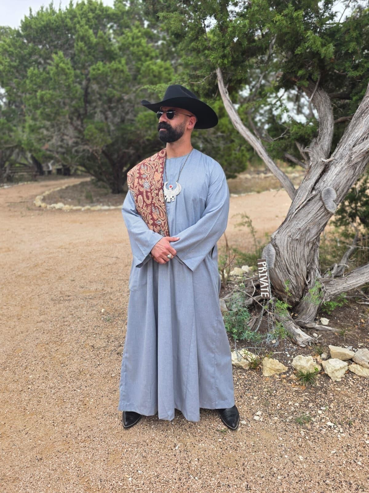 Kareem El-Ghayesh, standing on a dirt road by a tree, dons an ankle-length robe, cowboy boots, sunglasses and a Stetson hat.