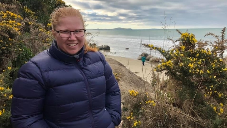 Rebecca Austin with glasses, smiling in a puffer jacket near white sand beach, yellow flower bushes around and cloudy sky.
