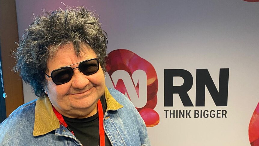 Richard Clapton wears sunglasses and smiles lightly at the camera, behind him is an RN backdrop