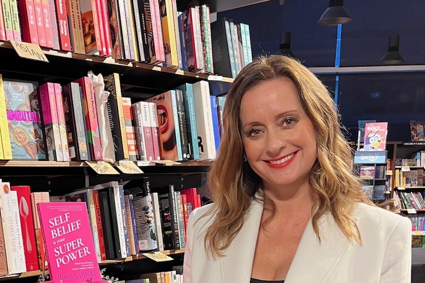 Tory Archbold wearing a white blazer standing next to pink books at a book store