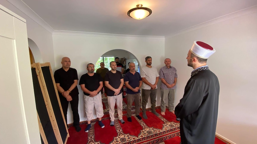 A Muslim man leading morning prayer with seven other man standing nearby in a white room.