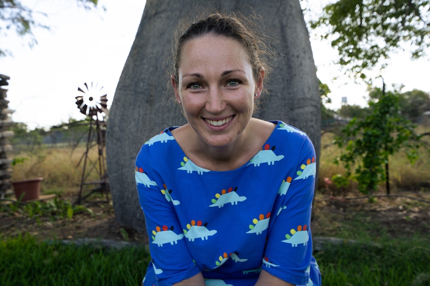 A smiling young woman sits in her backyard in outback Queensland with a thick tree trunk behind her.