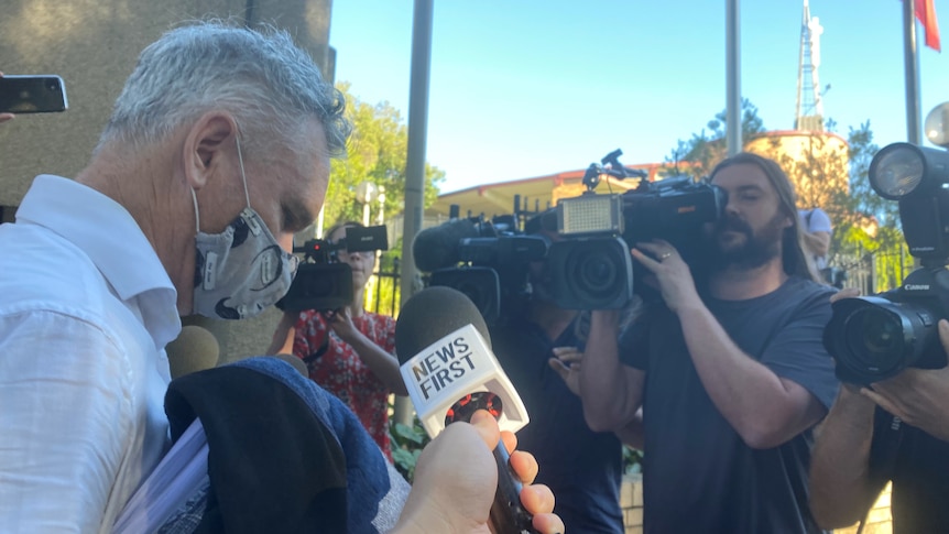 A man wearing a mask is confronted by the media