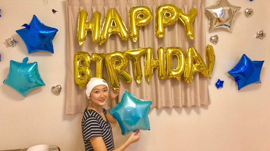 Rikako Ikee smiles while holding a star balloon in front of balloons spelling out "HAPPY BIRTHDAY". She has a bandaged head.