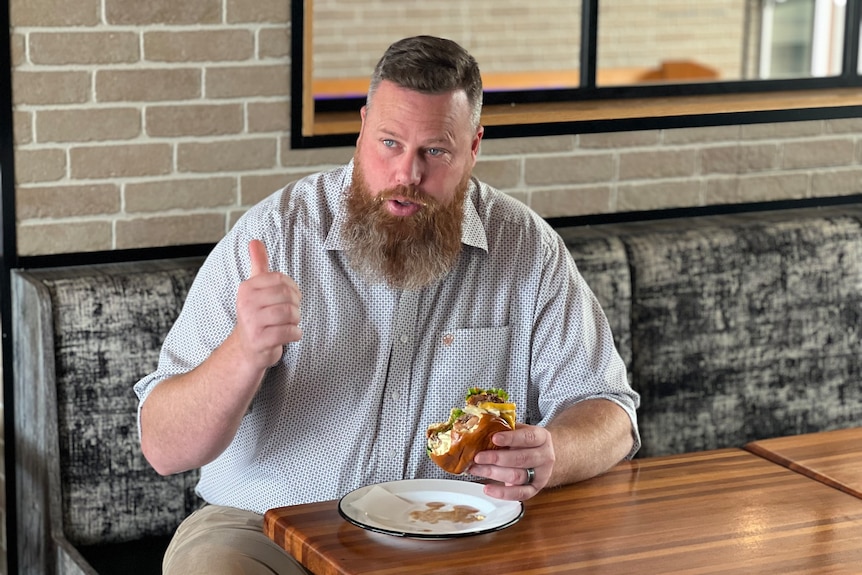 A man with a beard holding a burger in one hand and giving a 'thumbs up' with the other.