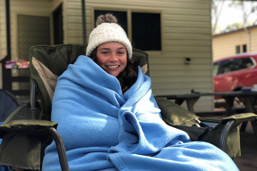 A woman smiles while sitting in a deck chair, wrapped tightly in a blue blanket