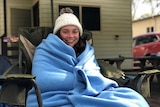 A woman smiles while sitting in a deck chair, wrapped tightly in a blue blanket