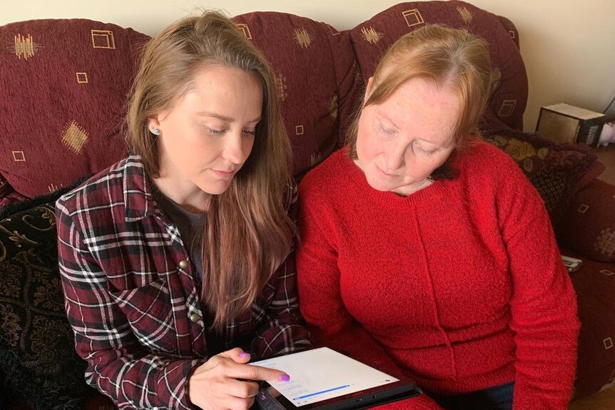 Two women look at a tablet while sitting on a couch.