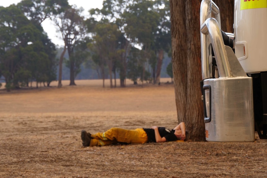 A firefighter lies on the ground in front of afire truck  with her arm over her face, head resting on a tree root, in a paddock.