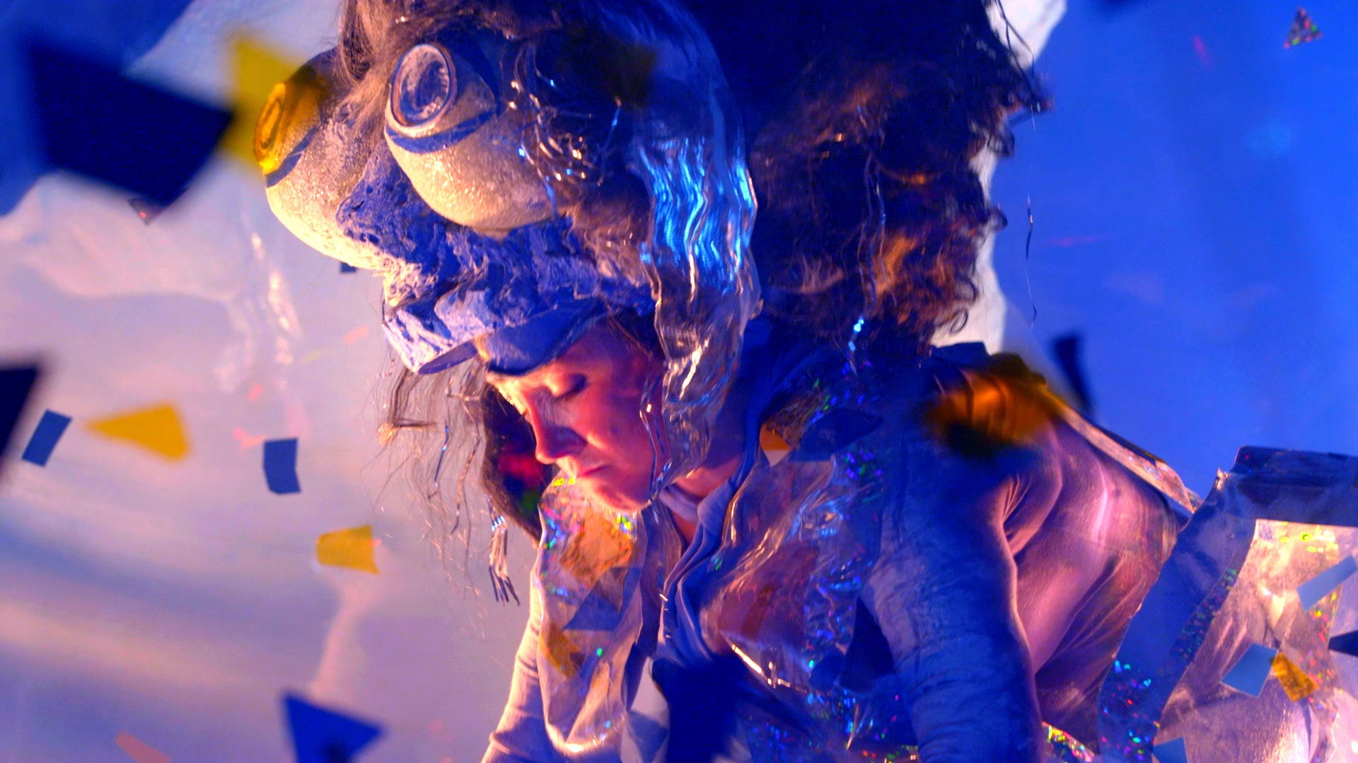 A still of a woman in an ostentatious costume, looking upset. She looks down, and her headpiece includes two upturned bowls.