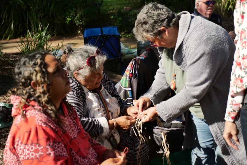 two women sit down with fibre in their hands, while another woman leans over helping them to weave