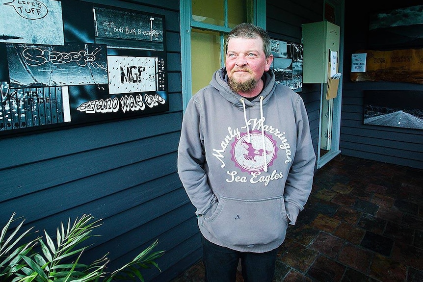 A man wearing a grey hoodie with pink writing stares off camera.