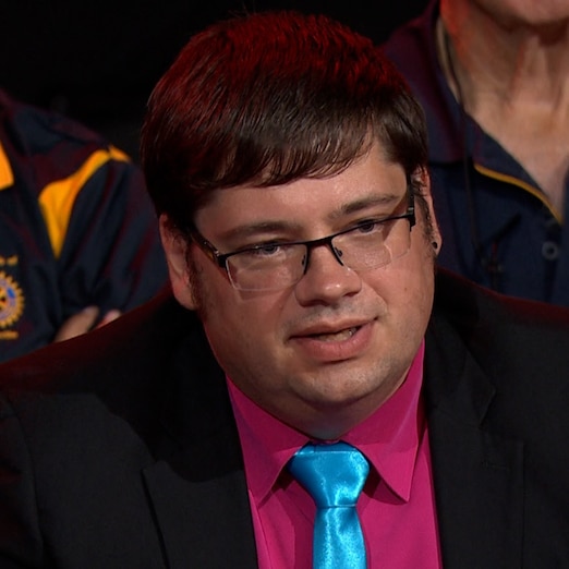 A person wearing glasses, a black jacket, maroon shirt and blue tie asks the panel a question