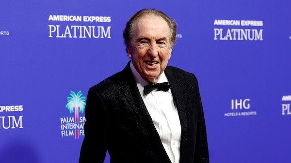 Eric Idle, a man in his 80s, wears a tuxedo on a red carpet, with a blue media wall behind him.