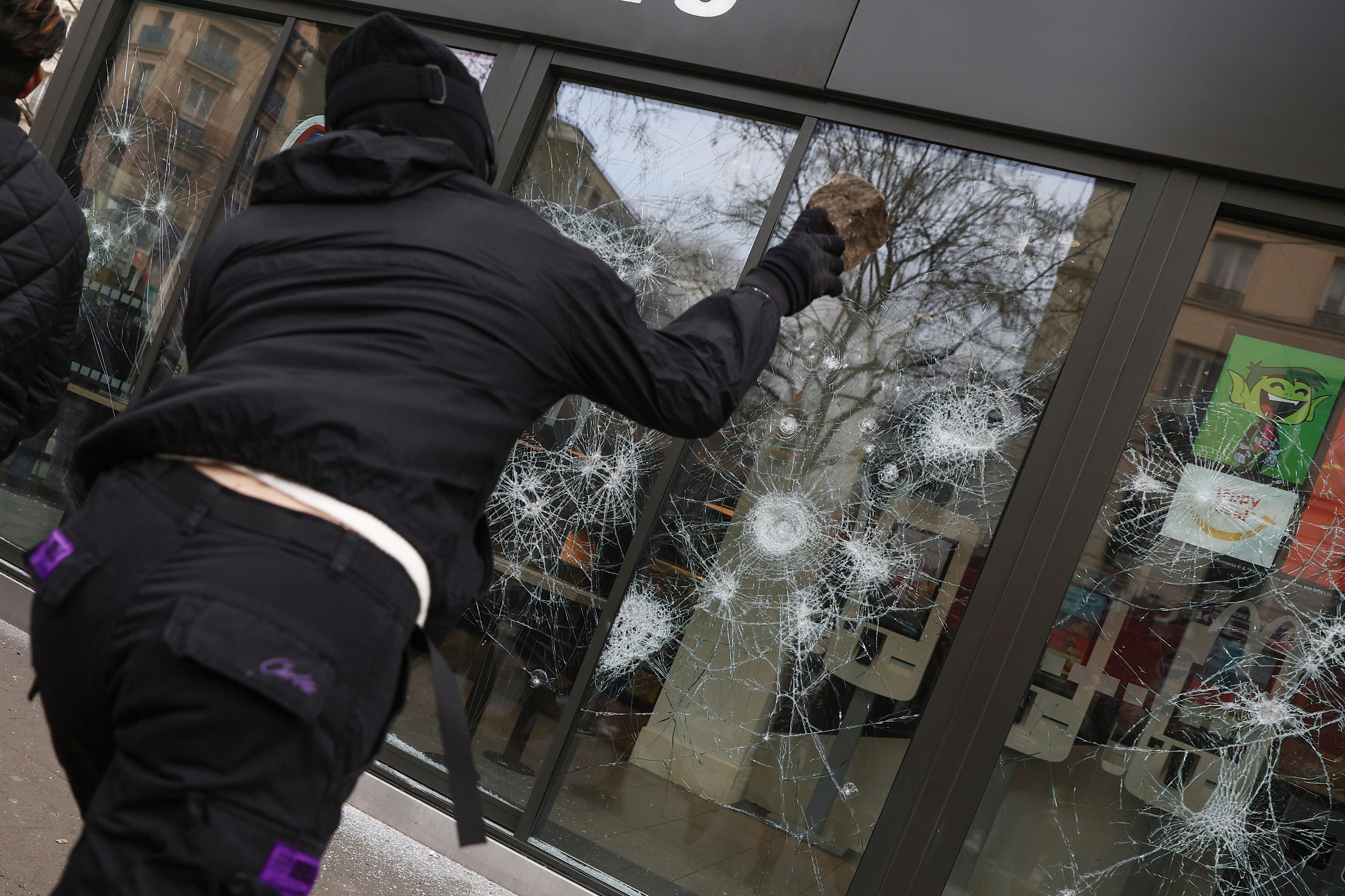 a person in a black outfit throws a rock at a window during protests in Paris