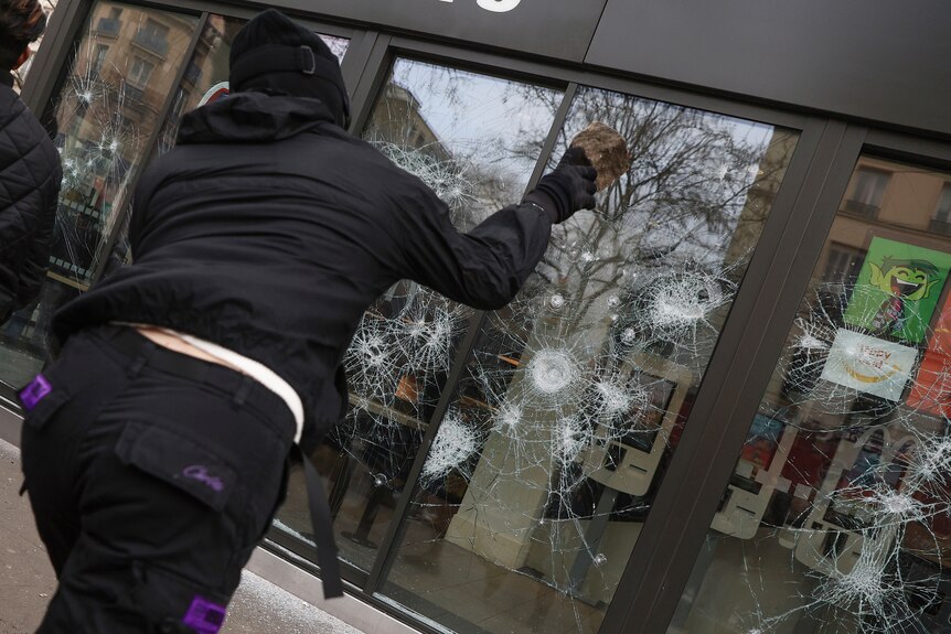 a person in a black outfit throws a rock at a window during protests in Paris