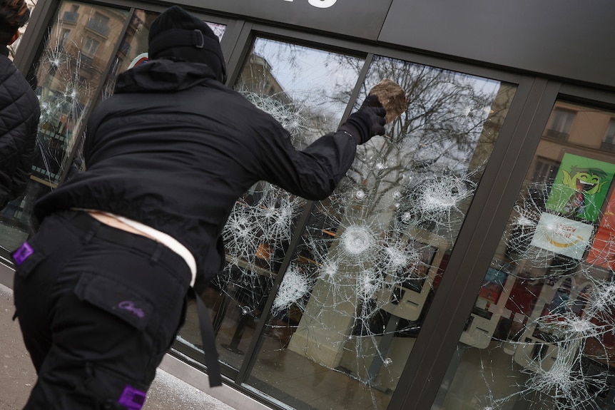 a person in a black outfit throws a rock at a window during protests in Paris