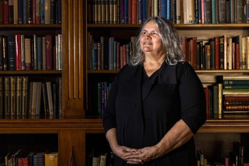 An Indigenous woman with shoulder-length grey hair poses for a portrait in front of shelves of books, smiling warmly.
