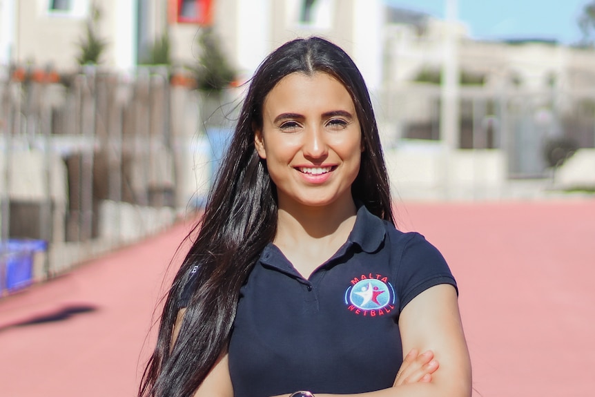 Nicole wears a navy polo with a Malta netball logo on one side, she stands next to a blurred background