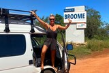 A woman standing on the doorframe of her van, next to a Welcome to Broome sign. 