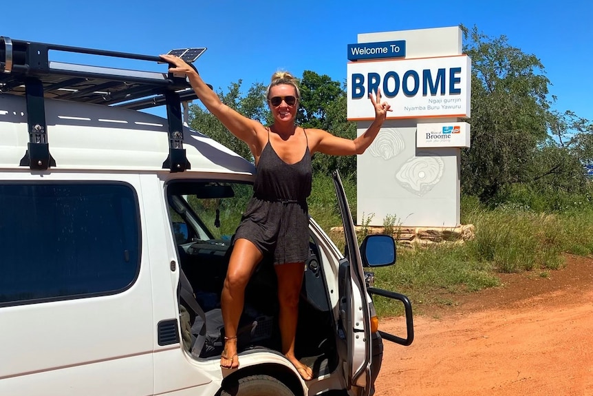 A woman standing on the doorframe of her van, next to a Welcome to Broome sign. 