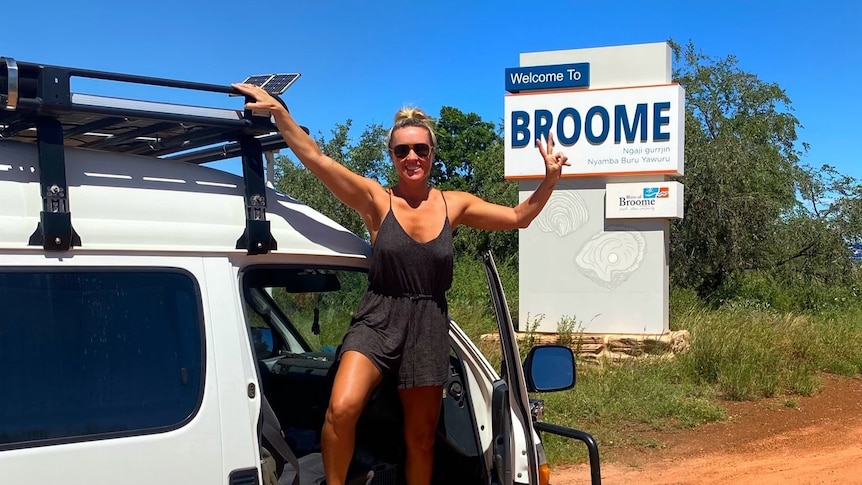 A woman standing on the doorframe of her van, next to a Welcome to Broome sign. 