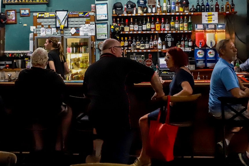 A group of men and women congregate at the bar of a pub, with alcohol and blue walls behind them