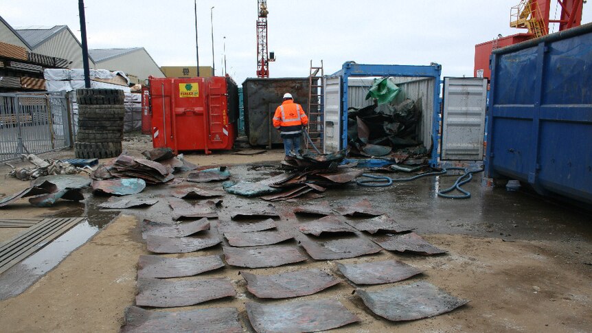 At an industrial site, you see rows of copper plates dating back to the medieval-era, with coloured shipping containers behind.